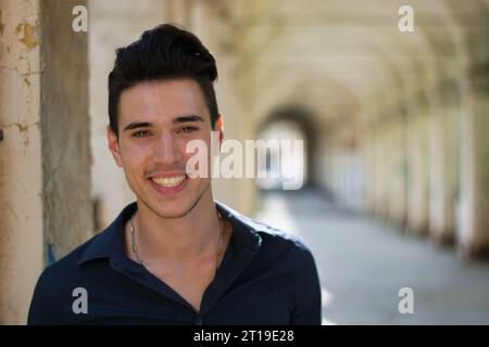 A young man is smiling for the camera. Photo of a young man with a bright smile, outdoor under a colonnade in European city. A cheerful young adult smiling with eye contact. Stock Photo
