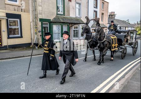 Funeral cortège with the funeral director leading a Victorian glass horse drawn hearse carriage with black horses and black plumage Stock Photo