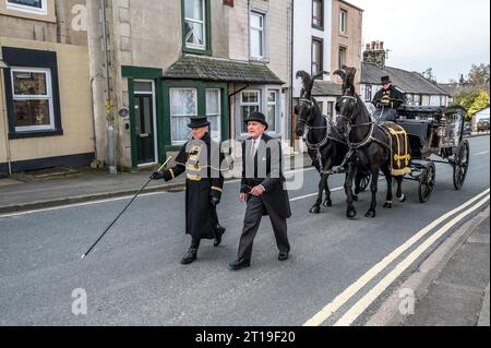 Funeral cortège with the funeral director leading a Victorian glass horse drawn hearse carriage with black horses and black plumage Stock Photo