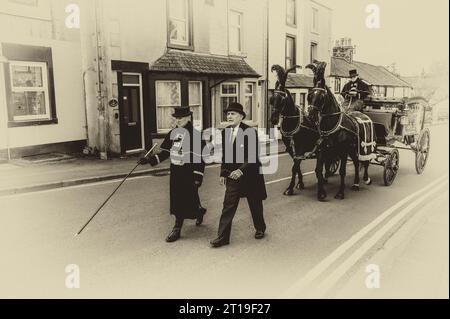 Funeral cortège with the funeral director leading a Victorian glass horse drawn hearse carriage with black horses and black plumage Stock Photo