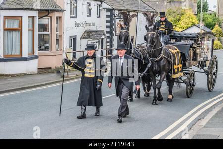 Funeral cortège with the funeral director leading a Victorian glass horse drawn hearse carriage with black horses and black plumage Stock Photo