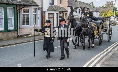 Funeral cortège with the funeral director leading a Victorian glass horse drawn hearse carriage with black horses and black plumage Stock Photo