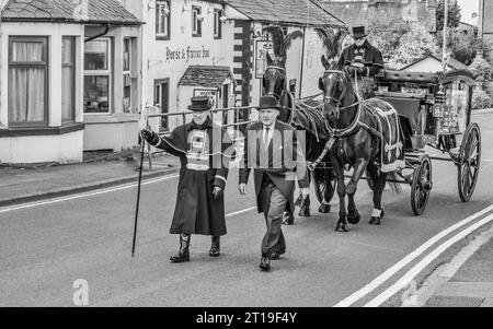 Funeral cortège with the funeral director leading a Victorian glass horse drawn hearse carriage with black horses and black plumage Stock Photo