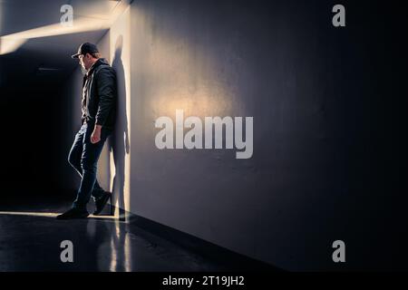 Serious sad man standing in dark corridor. Mystery guy in spotlight. Dramatic light and shadow. Stalker or outcast. Person waiting, hiding. Stock Photo
