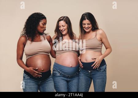 Diverse group of expecting mothers proudly display their baby bumps in a studio. Wearing jeans and bras, these young women, in their third trimester, Stock Photo