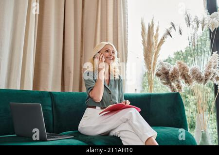 happy middle aged woman talking on smartphone and taking notes while sitting on sofa with laptop Stock Photo