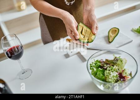 close up of cropped woman holding sliced avocado near lettuce in bowl and glass of red wine Stock Photo