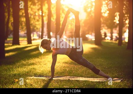 Beautiful sporty fit yogini woman practices yoga advanced side plank pose vasisthasana in the park at sunset Stock Photo