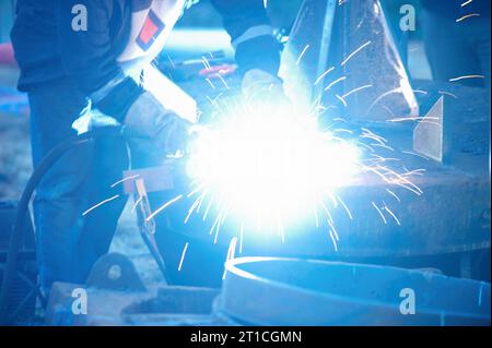 Industrial electrode welder with face shield and blue overall welding a steel pipe in workshop. Close up view. Stock Photo
