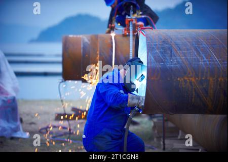 Industrial electrode welder with face shield and blue overall welding a steel pipe in workshop. side view. Stock Photo