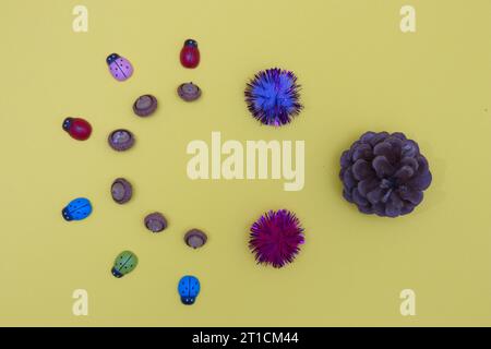 A cone against colorful ladybugs and acorns arranged in a semicircle on a yellow background. Flat lay minimalistic composition. Stock Photo