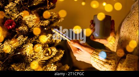 Side view of smartphone and cup in female hands in front of Christmas tree. Stock Photo