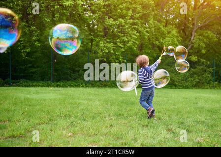 Outdoors portrait of cute preschool boy blowing soap bubbles on a green lawn at the playground Stock Photo