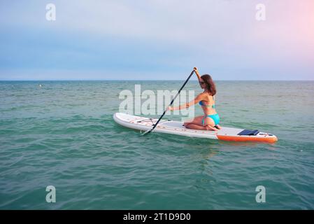 Womanl in a blue bikini sits on a sup board with a paddle in the sea against the sunny day Stock Photo