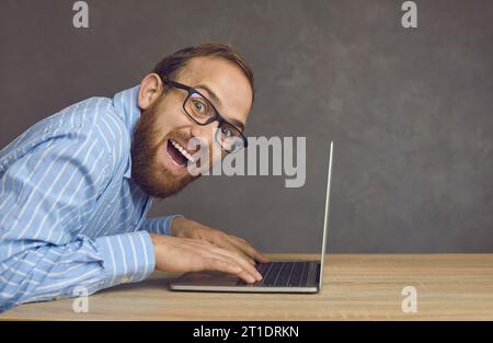 Side view portrait of a joyful crazy hacker or programmer sitting at a table typing on a laptop. Stock Photo
