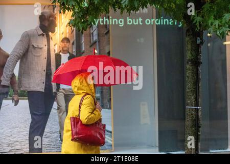 M&S Marks and Spencers 'Anything but Ordinary' Preston, Lancashire.  UK Weather. 13 Oct 2023.  Woman with red umbrella in yellow coat on a rainy start to the day in Preston. Stock Photo