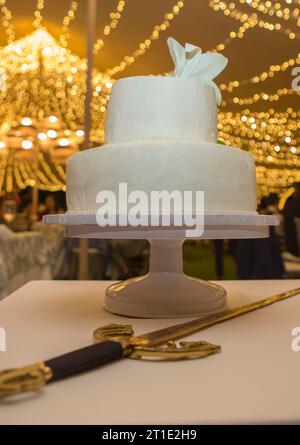 Fondant wedding cake over table with sword at banquet hall. Decorated tent with LED garlands as background Stock Photo