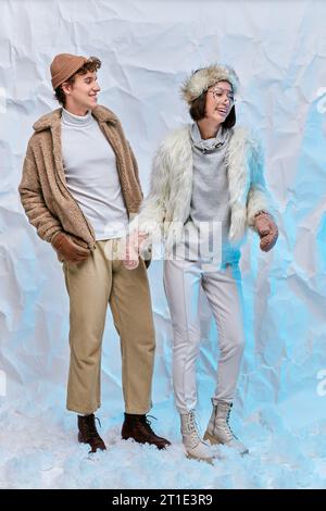 fashionable multiethnic couple in cold-weather attire laughing on snow in studio, winter happiness Stock Photo