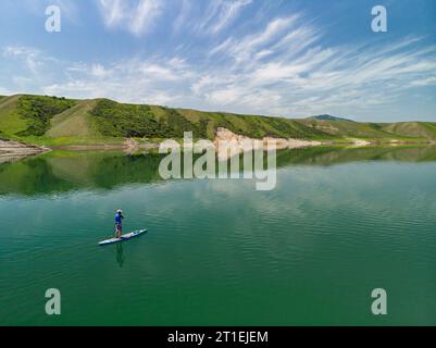 A bearded man swims on a SUP board on a lake among the rocks. Stand Up Paddling. Mountain lake with azure water Stock Photo
