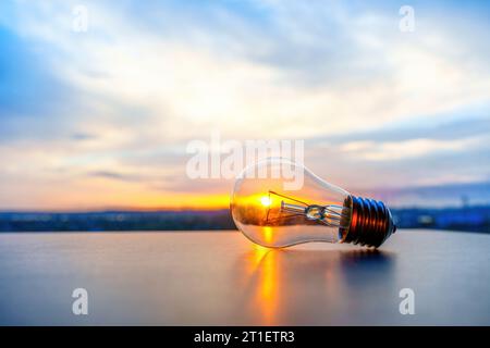 Detailed view of an incandescent light bulb with the sun setting behind the horizon in the background. Alternative energy sources and sustainability r Stock Photo