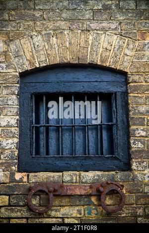 An ancient brick prison wall with a boarded up window with metal bars and rusty metal shackles beneath Stock Photo