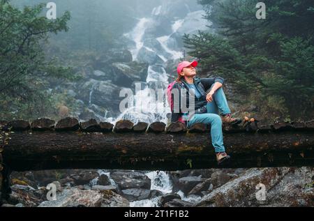 Young woman with backpack sitting on a wooden bridge and enjoying of power mountain river waterfall during Makalu Barun National Park trek in Nepal. M Stock Photo