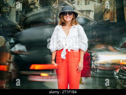 Cheerfully smiling female tourist standing on rush Nepalese narrow street inside the fast-moving town traffic with cars, motorcycles, and rickshaws. T Stock Photo
