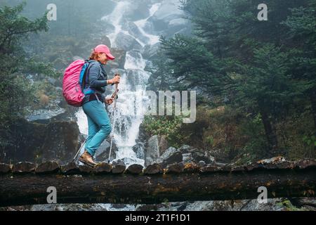 Young woman with backpack and trekking poles crossing wooden bridge near power mountain river waterfall during Makalu Barun National Park trek in Nepa Stock Photo