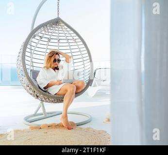 Beautiful middle-aged woman sitting in a comfortable hanging chair on the open house terrace with a modern laptop and browsing in internet Stock Photo