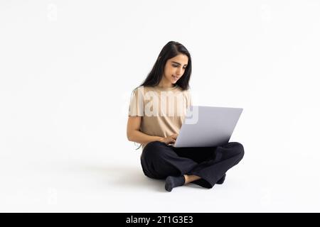 Online assistant, chatbot, artificial intelligence in daily life. Smiling beautiful young indian woman wearing casual outfit using laptop on white wal Stock Photo