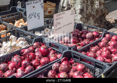 Red onions, garlic bulbs and white onions priced for sale in black plastic crates at the Saturday outdoor market in Arles, Provence, southern France. Stock Photo