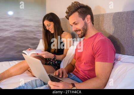 A Caucasian couple lounging on the hotel bed and looking at a computer and a book. Summer lifestyle Stock Photo