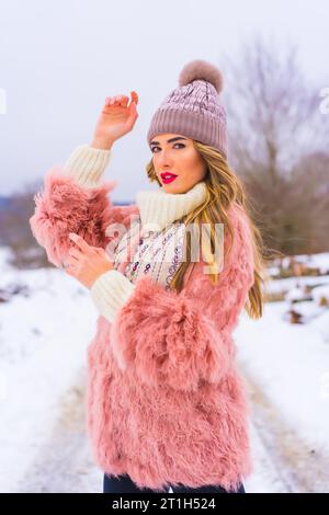 Portrait of young blonde model wearing pink fur jacket, winter boots and a purple hat in the snow. Lifestyle Stock Photo