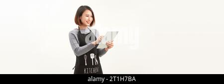Friendly cute female barista in black apron smiling at camera, using digital tablet to manage cafe orders Stock Photo
