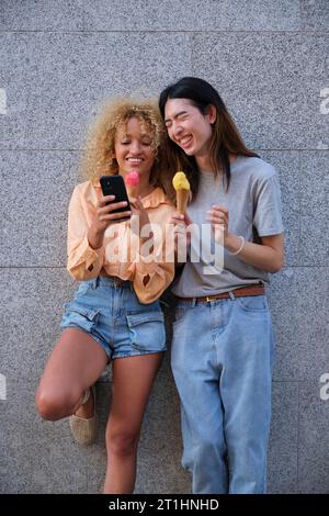 Two multiracial happy friends with ice creams using the smartphone in summer at street. Stock Photo