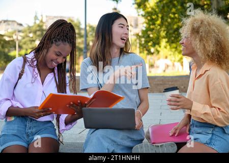 Three happy multiracial friends studying with a laptop and folder at a park. Stock Photo