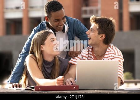 Cheerful group of people working together with a laptop in campus.Young multiracial friends studying and laughing outside using computer and notes. Stock Photo