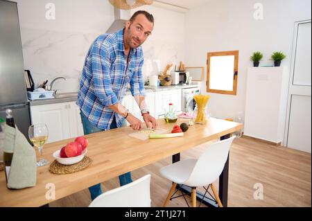 Young adult Caucasian handsome man in casual clothes, smiles looking at camera, cutting garlic, preparing a sauce for Italian pasta in the home kitche Stock Photo