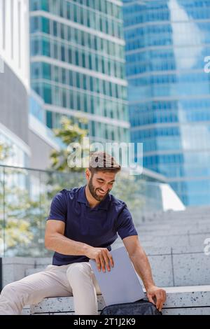 Smiling businessman putting laptop inside backpack while sitting on steps on skyscers background Stock Photo