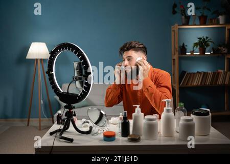 Male beauty blogger making video on smartphone set on ring lamp, showing facial cleans and advertising skincare products. Blogging career Stock Photo