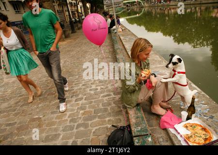 A woman is sharing a pizza with her pet dog on the side of Canal St. Martin in Paris, France Stock Photo