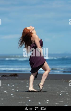 Redhead woman in purple brown long dress standing on sandy beach with head looking upward and eyes closed, sunbathing on background blue sea on summer Stock Photo