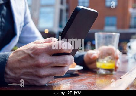Close-up of unrecognizable Caucasian adult man, sitting at outdoor restaurant, holding phone sending messages with one hand while having sandwich with Stock Photo