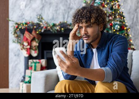 Upset sad and disappointed man received message online about bad news, hispanic sitting on sofa in living room, using app on smartphone, depressed unhappy reading internet pages. Stock Photo