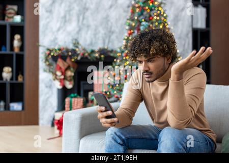 Upset sad and disappointed man received message online about bad news, hispanic sitting on sofa in living room, using app on smartphone, depressed unhappy reading internet pages. Stock Photo