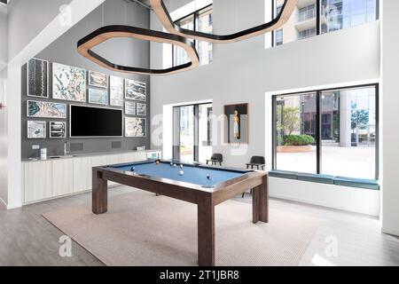 A modern billiards room with a pool table with a blue top, a modern light fixture hanging from the tall ceilings, and TV mounted on the wall. Stock Photo