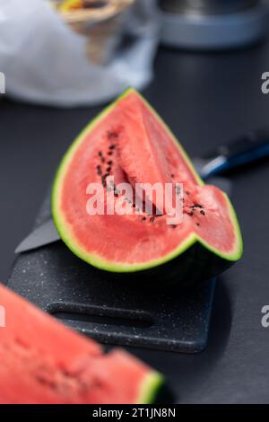 Vertical portrait of a quarter slice of a green cut watermelon lying on a plastic cutting board next to a knife. The piece of the fruit is ready to be Stock Photo