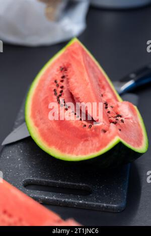 A portrait of a quarter slice of a green cut watermelon lying on a plastic cutting board next to a knife. The piece of the fruit is ready to be eaten Stock Photo