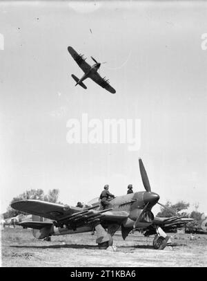 Royal Air Force- 2nd Tactical Air Force, 1943-1945. A Hawker Typhoon half-rolls over Thorney Island, Hampshire, after taking off to attack enemy communications targets on the French coast, watched by ground crew working on Typhoons in a dispersal on the airfield. Stock Photo