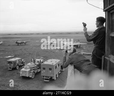 Royal Air Force- Italy,the Balkans and South-east Europe, 1942-1945. A view from the watch office at Foggia Main, Italy, as an airman prepares to fire a Very pistol from the balcony. Parked immediately below the tower are two airfield ambulances and a Crossley Fire Crash Tender. Beyond the vehicles are a number of visiting aircraft, including (left to right): a Lockheed P-38 of the 15th USAAF; two communications aircraft, a Fairchild Argus of the Middle East Communications Squadron and a Vultee Stinson Vigilant, and De Havilland Mosquito NF Mark XIII, MM567 'RO-A', of No. 29 Squadron RAF, unde Stock Photo